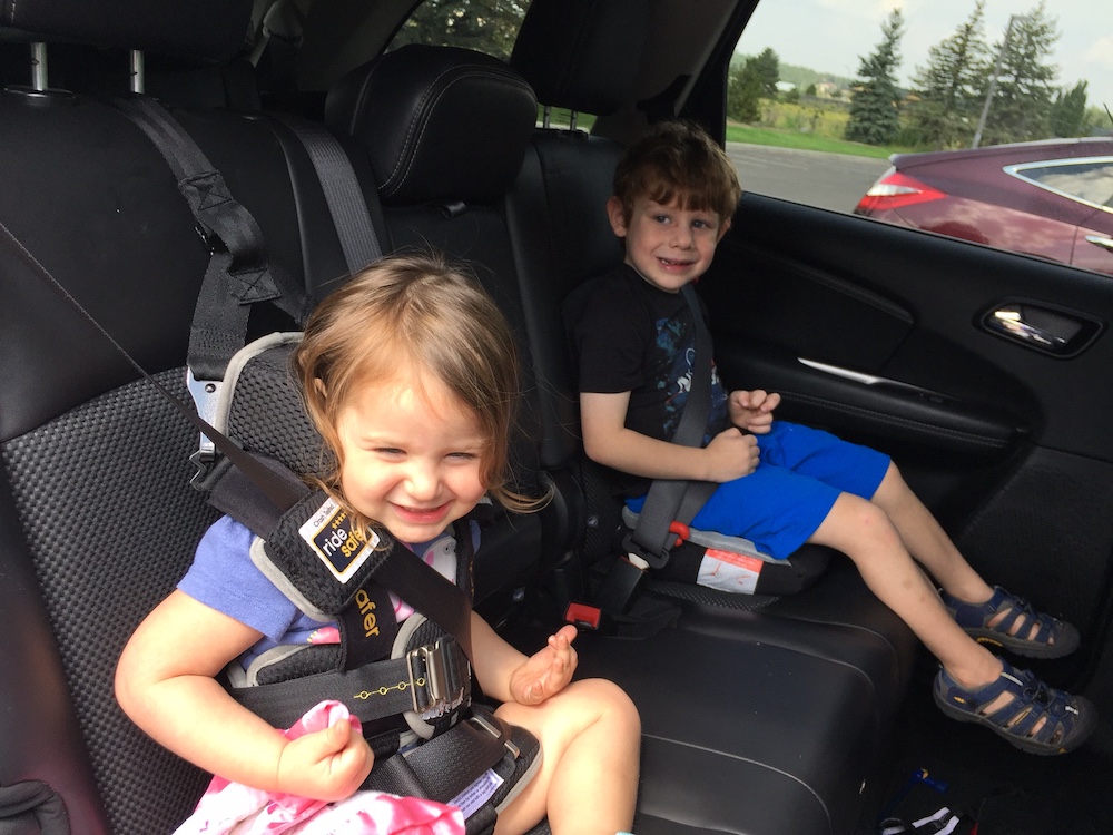 folding car seat for travel: young girl in foreground in Ride Safer travel vest, young boy in background sitting on Bubblebum inflatable booster seat