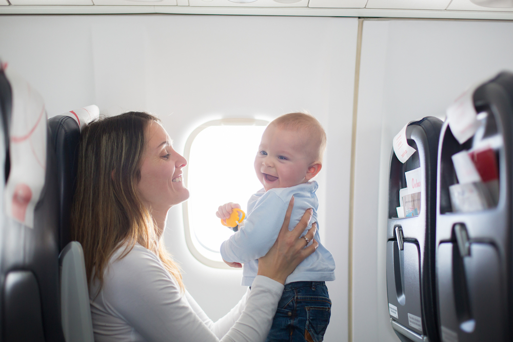 Travel Car Seat Mom - A woman holding a baby on an airplane with a car seat.