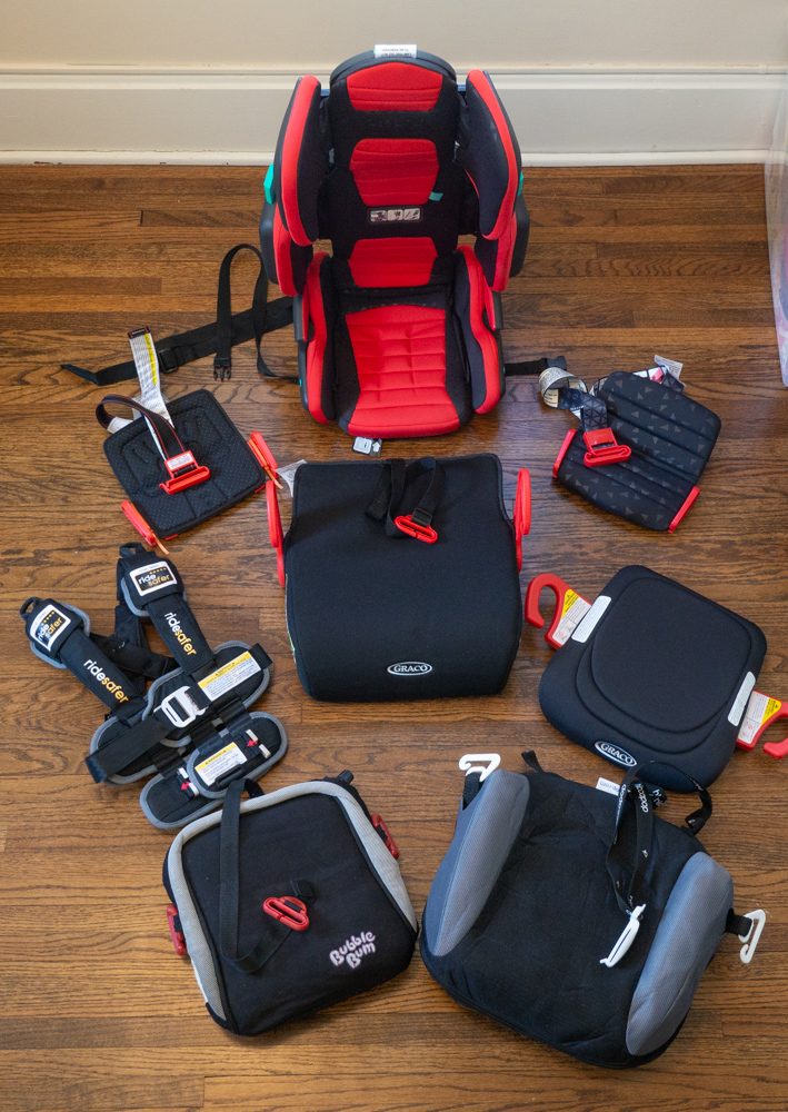 Best travel booster seat options unfolded and ready to use in a circle on a wood floor. Center: Graco Turbo GO; Clockwise from top: hifold, mifold comfort, Graco RightGuide, hiccapop Uberbooster, Bubblebum, Ride Safer travel vest, mifold original